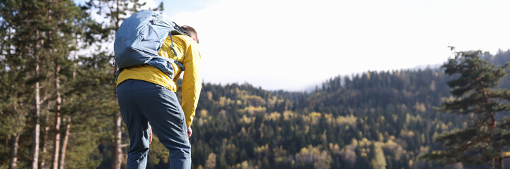 Tired male traveler with heavy backpack bending over near forest lake back view