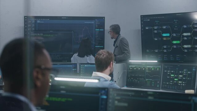 Mature big data scientist with female colleague analyze real-time charts displayed on multiple big screens. Technical support specialists work at computers using professional software. Cyber security.
