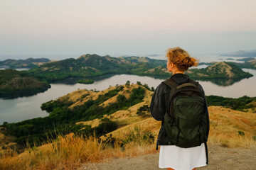 A young girl, blonde, with a backpack, stands on top of a mountain, rear view, overlooking a beautiful panorama of the islands and bays in Indonesia.