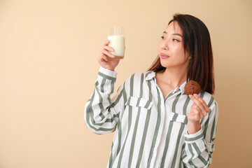 Young Asian woman with milk and cookie on beige background
