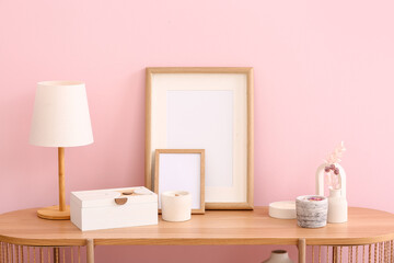 Holders with candles, blank frames, lamp and decor on table near pink wall in room