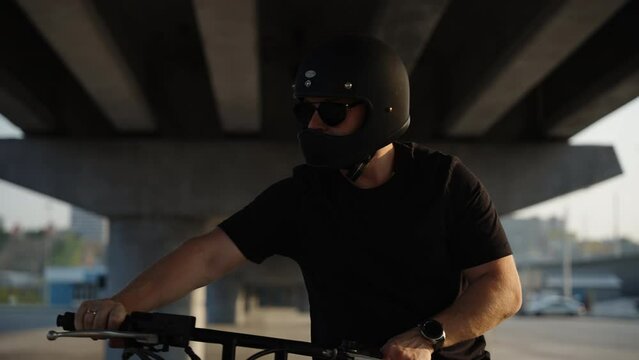 A biker holds the motorcycle by the handlebars and walks on the asphalt with the bike muffled. The biker has a black helmet with glasses. A serious biker on a motorcycle walks under a road bridge. A