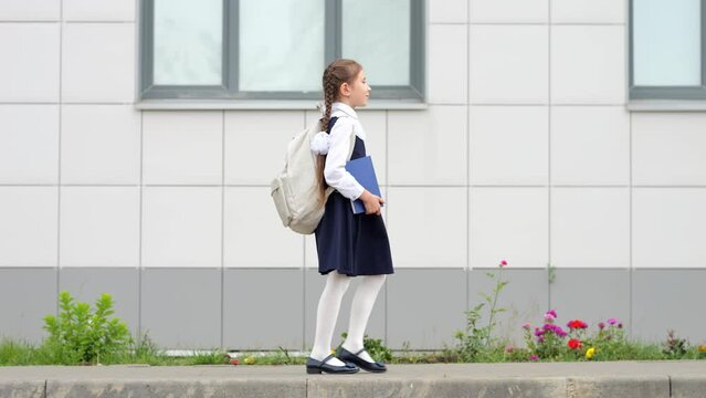 Schoolgirl with long hair carrying white backpack and blue book goes to modern school. Elementary student with braids starts new day at school, side view