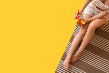 Young beautiful woman with tasty cocktail sitting on beach blanket against yellow background....