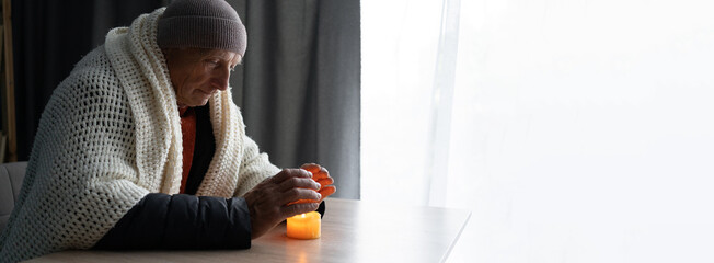 Tired old man warm hands over candle light at home no heating. Shutdown of heating and electricity,...