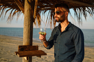 Contemplative Man with Wine on the Beach - A bearded man in sunglasses reflects with a glass of wine, under the filtered sunlight of a straw umbrella.