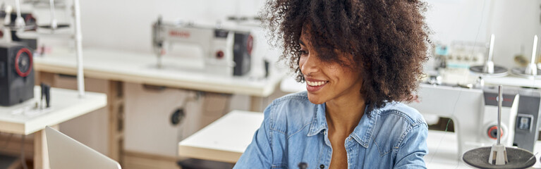 Positive young African-American female tailor watches video lesson via modern laptop sitting at workplace