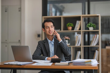 Obraz premium Portrait of a professional Asian businessman looking some details on a paper while talking on the phone with his supplier in the office.