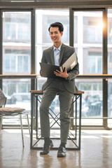 portrait of a handsome and successful Asian businessman or male banker in a formal business suit stands in his office with a cup of coffee and a document folder in his hand.