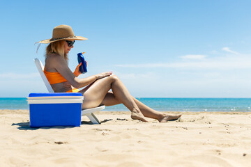beach background with girl, slender girl in a swimsuit sits on a sun lounger smears her legs with sunblock, a blue portable refrigerator for drinks stands nearby. the concept of relaxing on the beach