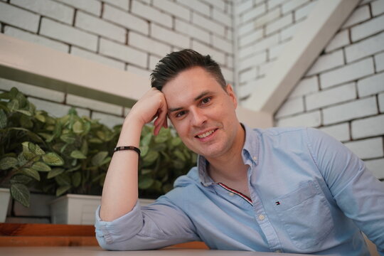 Casual Portrait: A young man with a stylish haircut and a blue collared shirt leans his head on his hand, smiling warmly at the camera in a relaxed indoor setting.