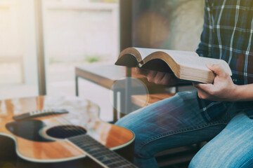 Close up of a man hands hold and reading the holy bible while sitting on wooden bench in the room with a guitar on wooden table, christian bible study, praise and worship concept with copy space