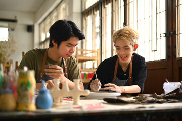 Cheerful young asian gay couple painting ceramics, creating handmade earthenware in workshop