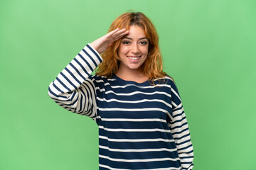 Young caucasian woman isolated on green screen chroma key background saluting with hand with happy expression