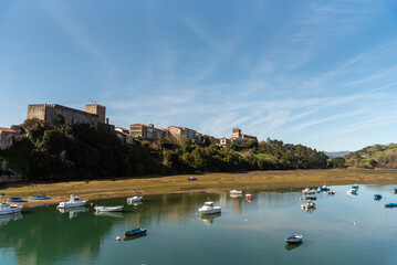 View of the bay of the tourist town of San Vicente de la Barquera in Cantabria at low tide, surrounded by vegetation and many colourful boats on its crystal clear waters during the sunset
