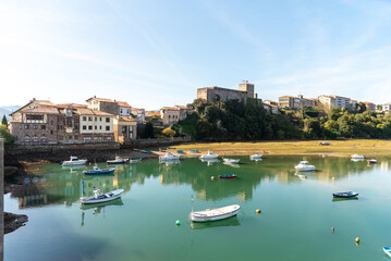 Fototapeta na wymiar Bay of the tourist village of San Vicente de la Barquera in Cantabria at low tide, surrounded by vegetation and many colourful boats on its crystal clear waters during the sunset on a summer day