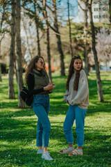 Fototapeta premium Full length photo of two lovely girls standing on a grass in the park. Brunette girl was distracted by something while talking to her friend