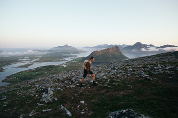 Healthy fit man with lightweight backpack hike up tall mountain in Lofoten archipelago in Norway. Amazing scenery with mountains and ocean in background. Hiking during midsummer midnight sun - obrazy, fototapety, plakaty