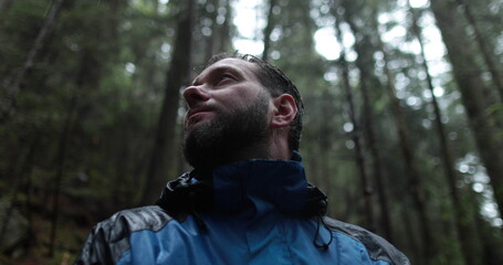 Close-up portrait of a young bearded man sitting in nature between forest trees and looking around.