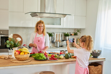 Daughter photographing mother by phone while preparing food in kitchen at home