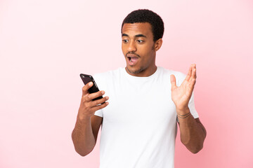 African American handsome man on isolated pink background looking at the camera while using the mobile with surprised expression