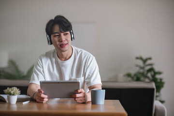 A focused young Asian male college student or freelancer using his digital tablet at a table in his living room.