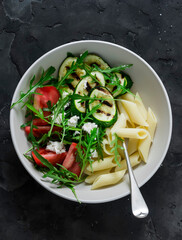 Vegetarian lunch - pasta and grilled zucchini, tomatoes, arugula, feta salad on a dark background, top view
