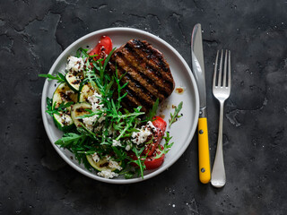 Steak and summer vegetable salad with grilled zucchini, tomatoes and arugula on a dark background, top view