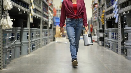 Woman shown from waist down, carrying can of paint and roller walking down paint aisle in hardware...