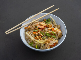 Shrimps and noodles, vegetables and peanuts in a white bowl with 2 chopsticks on top.