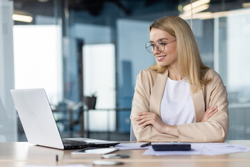 Obraz premium A young businesswoman is sitting in the office at the desk in front of the laptop, looking at the monitor with a smile. Participates in business training, interviews, business meetings via video call