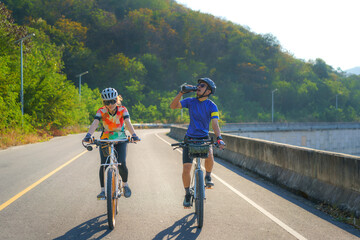 Fototapeta premium Tired Asian cyclist man drink water from a bottle while riding together for exercise around the lake in the morning