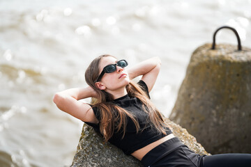 A beautiful young girl in sunglasses is lying on a rock on the seashore