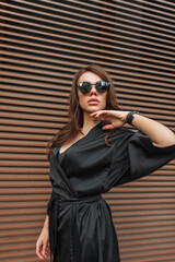 Beautiful fashion young glamour chic model girl with cool sunglasses in a stylish black dress stands and poses near a rusty metal wall