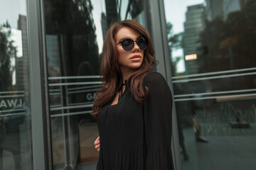 Elegant beautiful young fashionable gorgeous girl model with brunette hairstyle with cool vintage sunglasses in black trendy fashion dress in the city