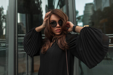 Fashion female street portrait of beautiful stylish glamorous cool woman with vintage sunglasses in fashionable black dress near modern glass building. Pretty lady