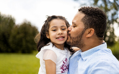 Fototapeta premium A dark-skinned father poses with his daughter in a park at sunset. The African adult gives a kiss on the face to the little girl looking at the camera. Concept of African-American parents.