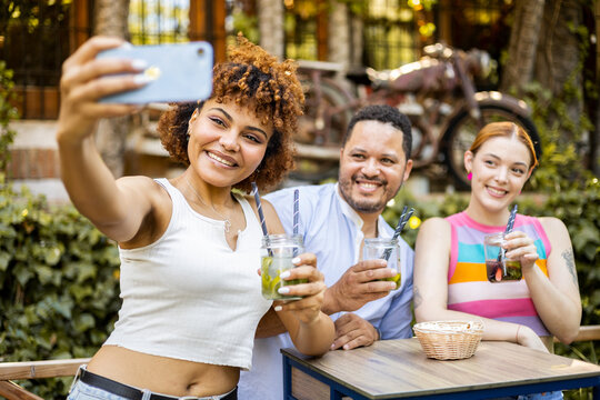 A group of 3 young multi-ethnic people take a picture with the mobile phone in a restaurant with vegetation while drinking mojitos. Concept of parties with cocktails, combined with alcohol.