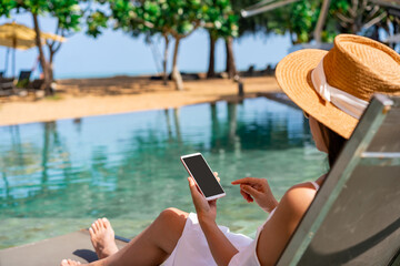 Young woman traveler relaxing and using a mobile phone by a resort pool while traveling for summer...