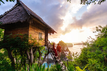 Young couple traveler enjoying and looking beautiful sunrise at the tree house in Nusa Penida island Bali, Indonesia - obrazy, fototapety, plakaty