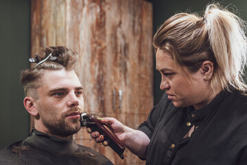 Young unshaven man in barbershop, woman hairdresser trims beard to client