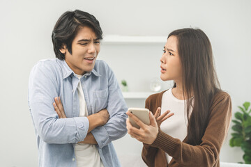 Infidelity, suspicion asian young couple love fight relationship, wife holding cellphone, smartphone cheating on phone, scolding husband about mistrust, distrust and jealousy when sitting at home.