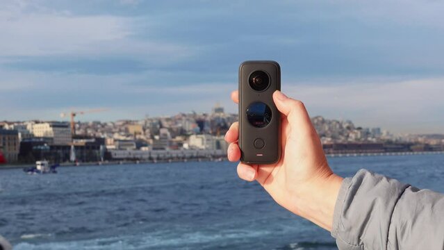 Hand of photographer, stoker holds action camera 360 shoots blog about trip video of sea trip, picturesque urban coastal landscape. Tourist shoots reportage about ship excursion on panoramic shooting