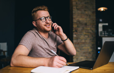 Skilled male blogger in optical spectacles for vision protection using cell application for online phoning in coworking space, Caucaisan man with laptop and textbook calling via smartphone