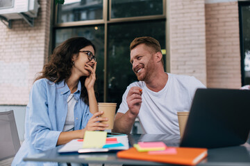 Happy startupers with modern laptop and paperwork laughing and rejoicing during cooperation meeting in street cafe, cheerful Caucasian man and woman enjoying positive mood for studying