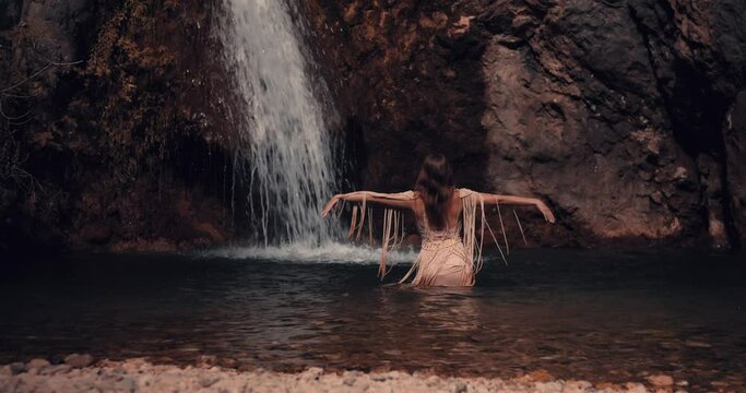 A young girl enters the water, stops in front of the waterfall and raises her hands up