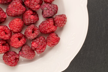 Several sweet raspberry berries with white ceramic plate on slate stone, macro, top view.