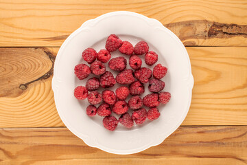 Several sweet raspberry berries with white ceramic plate on wooden table, macro, top view.