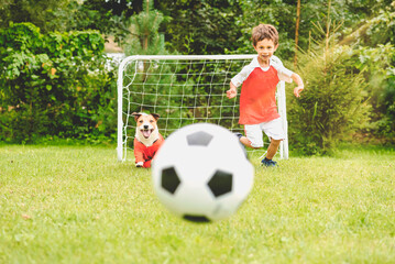 Happy kid in soccer kit playing football with family pet dog at garden lawn on summer day