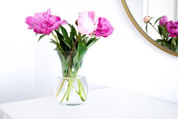 Pink peonies in the apartment. Home decoration, pink peonies on table in white room. Vase with beautiful peony flowers on table in bedroom. Flowers in a vase at home. Mirror reflection, gold frame